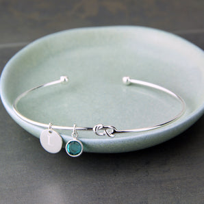 Silver bangle with initial disc charm and blue zircon birthstone shown resting in a dish.