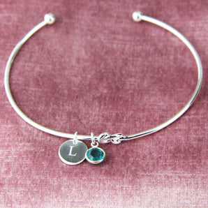 Silver bangle with initial disc charm and blue zircon birthstone shown against a pink velvet background
