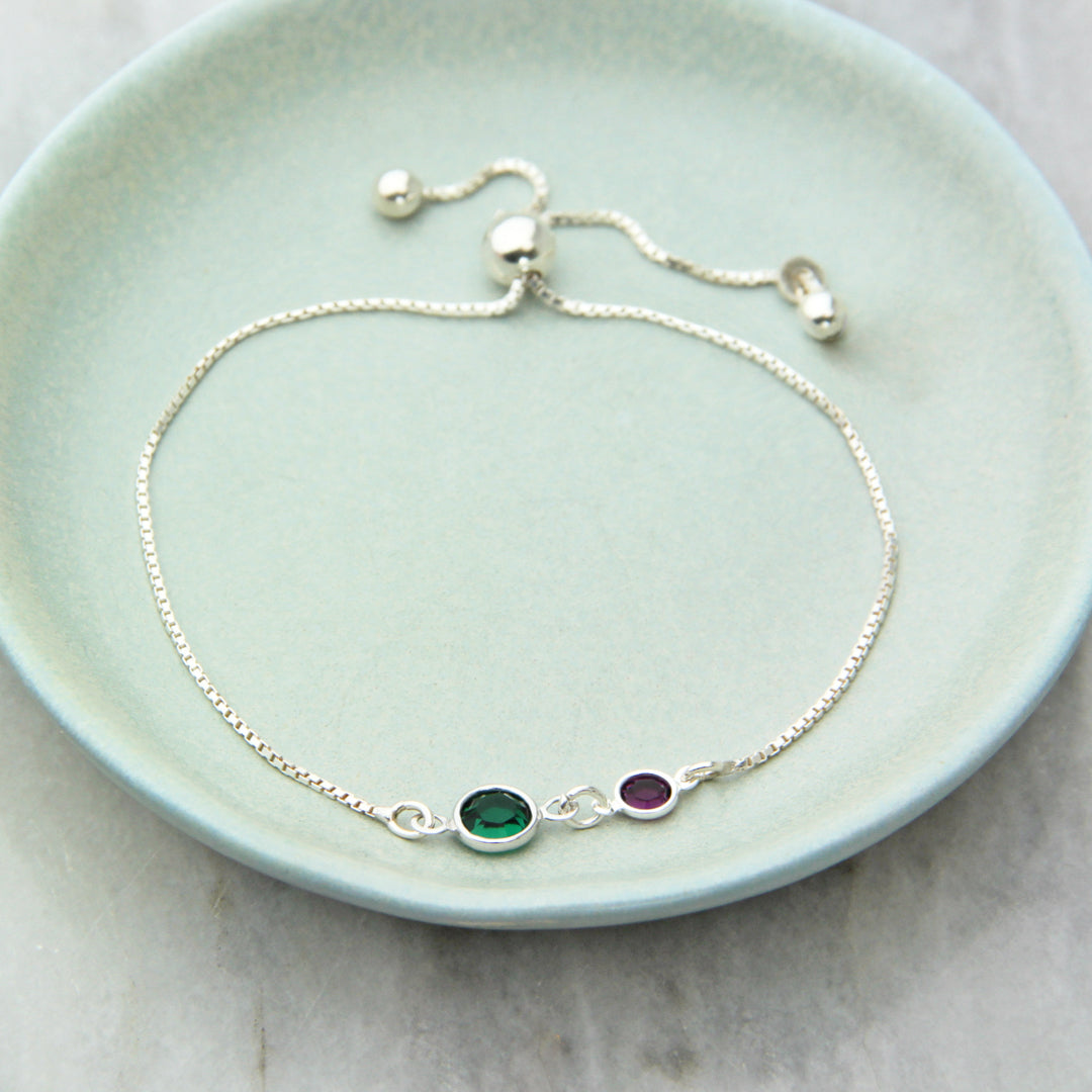 Adjustable silver bracelet with emerald and amethyst birthstones displayed in a dish